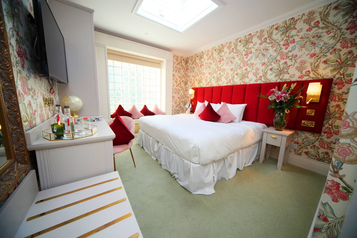 A bedroom with a large bed, a bedside table, a desk with a globe and candles, and a window with a view.