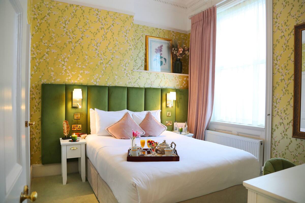 A hotel room with a green upholstered headboard, white bedding, a bedside table with a lamp, and a tray with breakfast items.