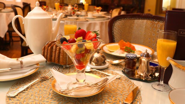 Una escena de desayuno con un bol de fruta, tetera y diversos productos para el desayuno sobre una mesa.