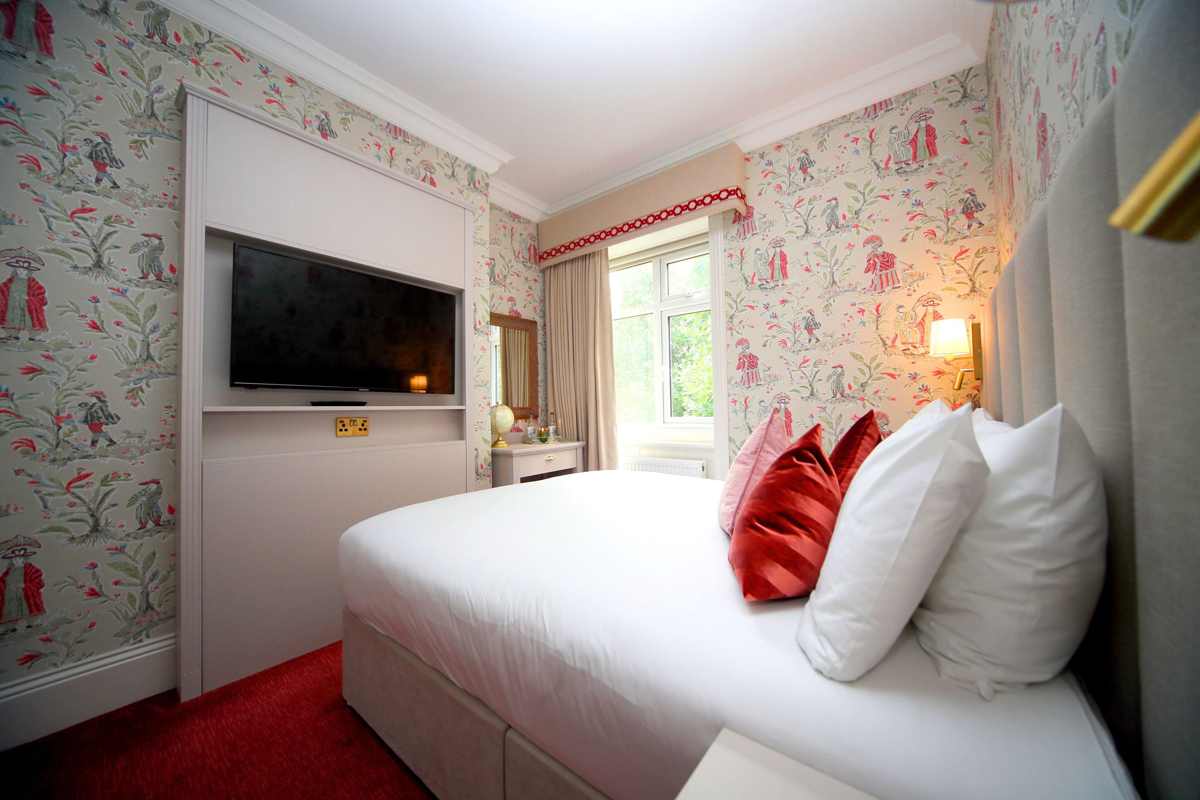 A hotel room with floral wallpaper, a bed with white and red pillows, a television on the wall, and a window with curtains.
