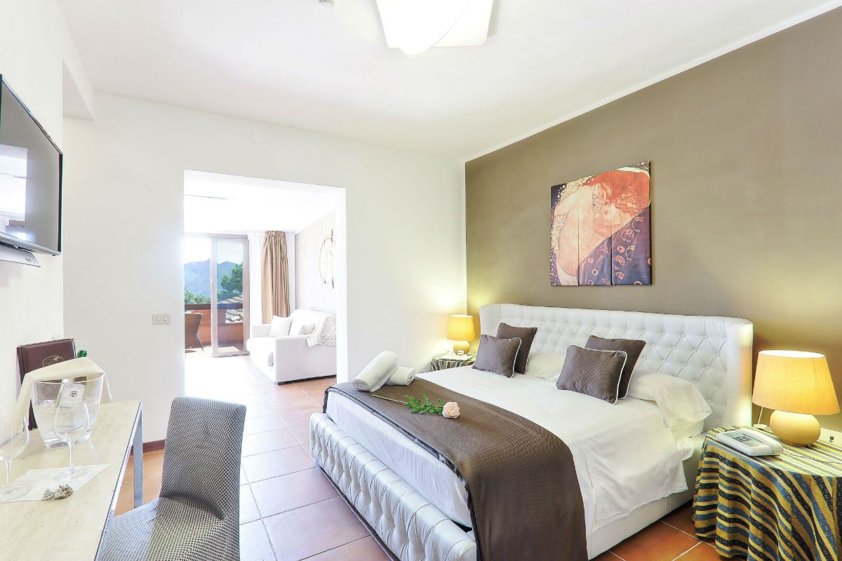 Quarto de hotel com cama, mesas de cabeceira, televisão, área de refeições e vista para o exterior.