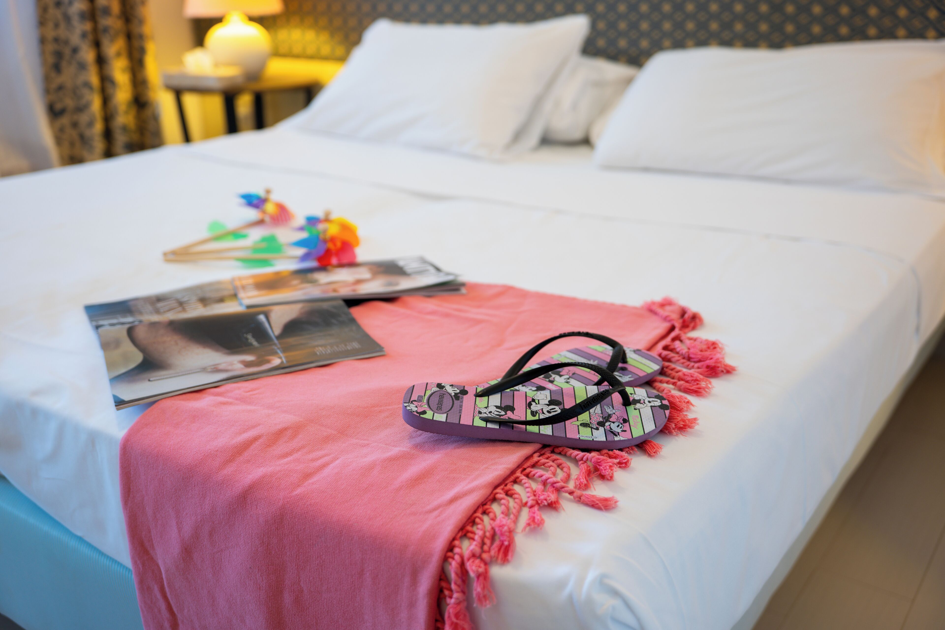 Un letto con una coperta rosa, un paio di ciabatte e alcune riviste.
