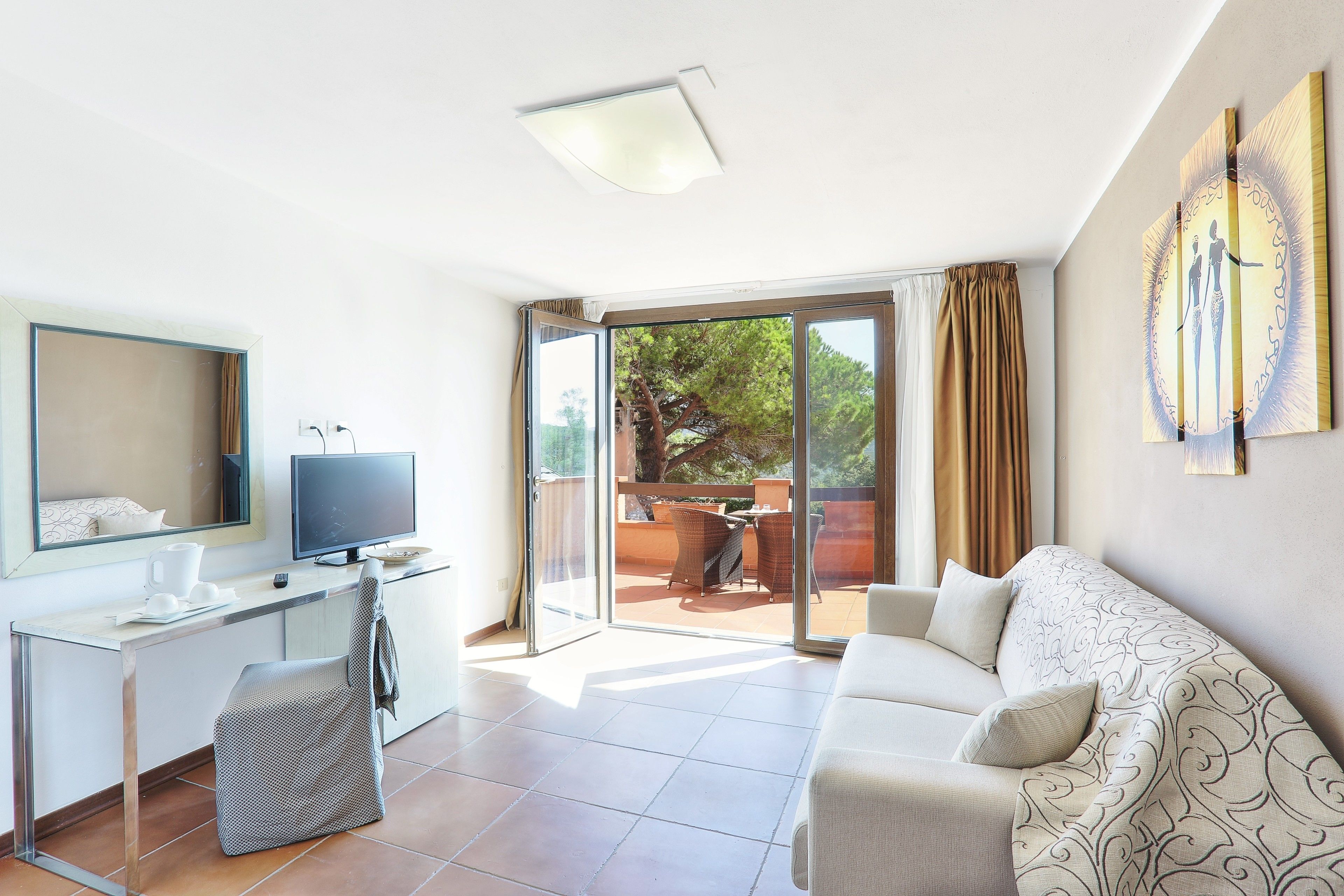 A modern hotel room with a desk, a flat-screen TV, a sofa, and a sliding glass door leading to an outdoor patio.
