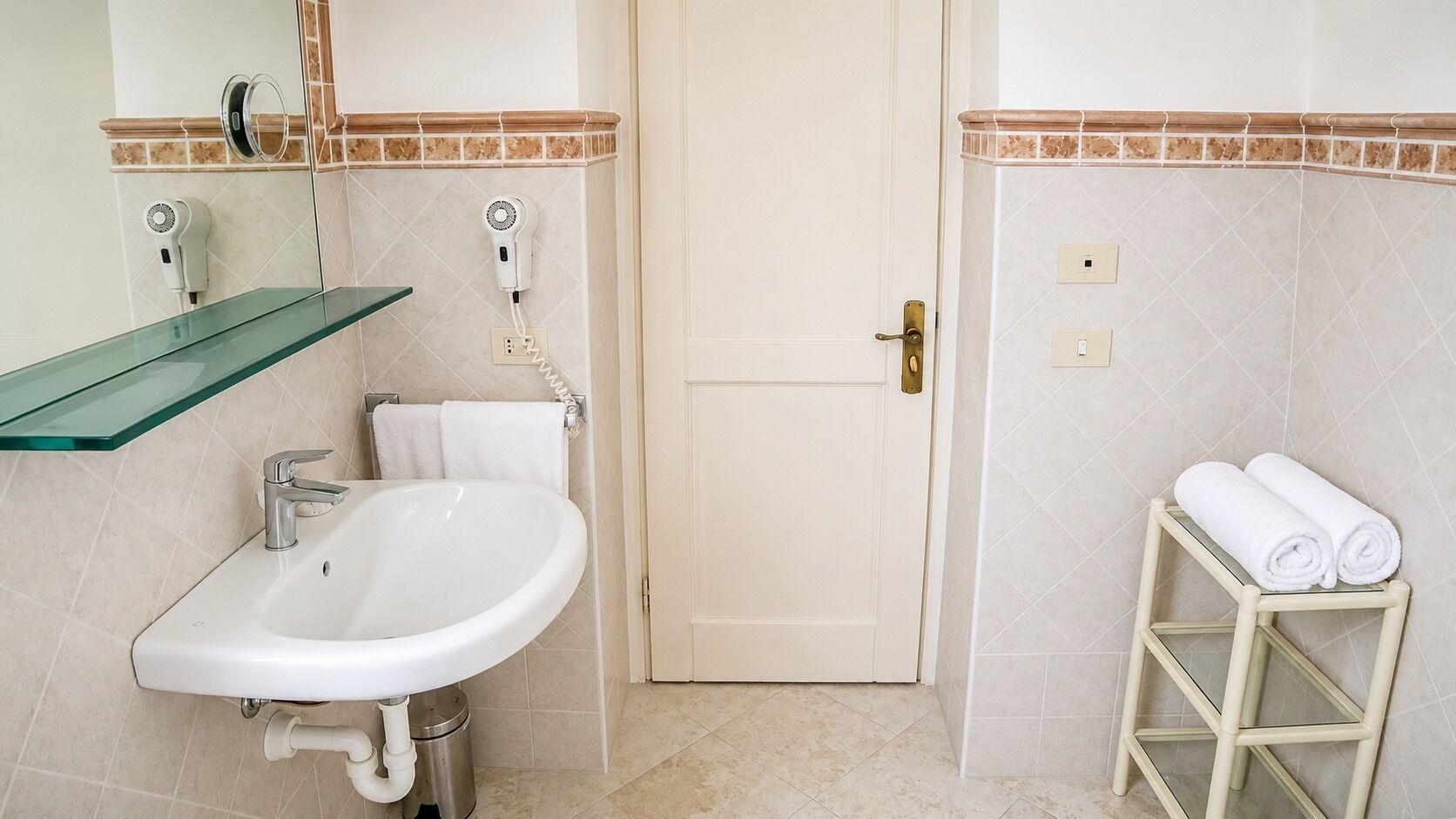A bathroom with a white sink, a glass shelf, a towel rack, and a door.
