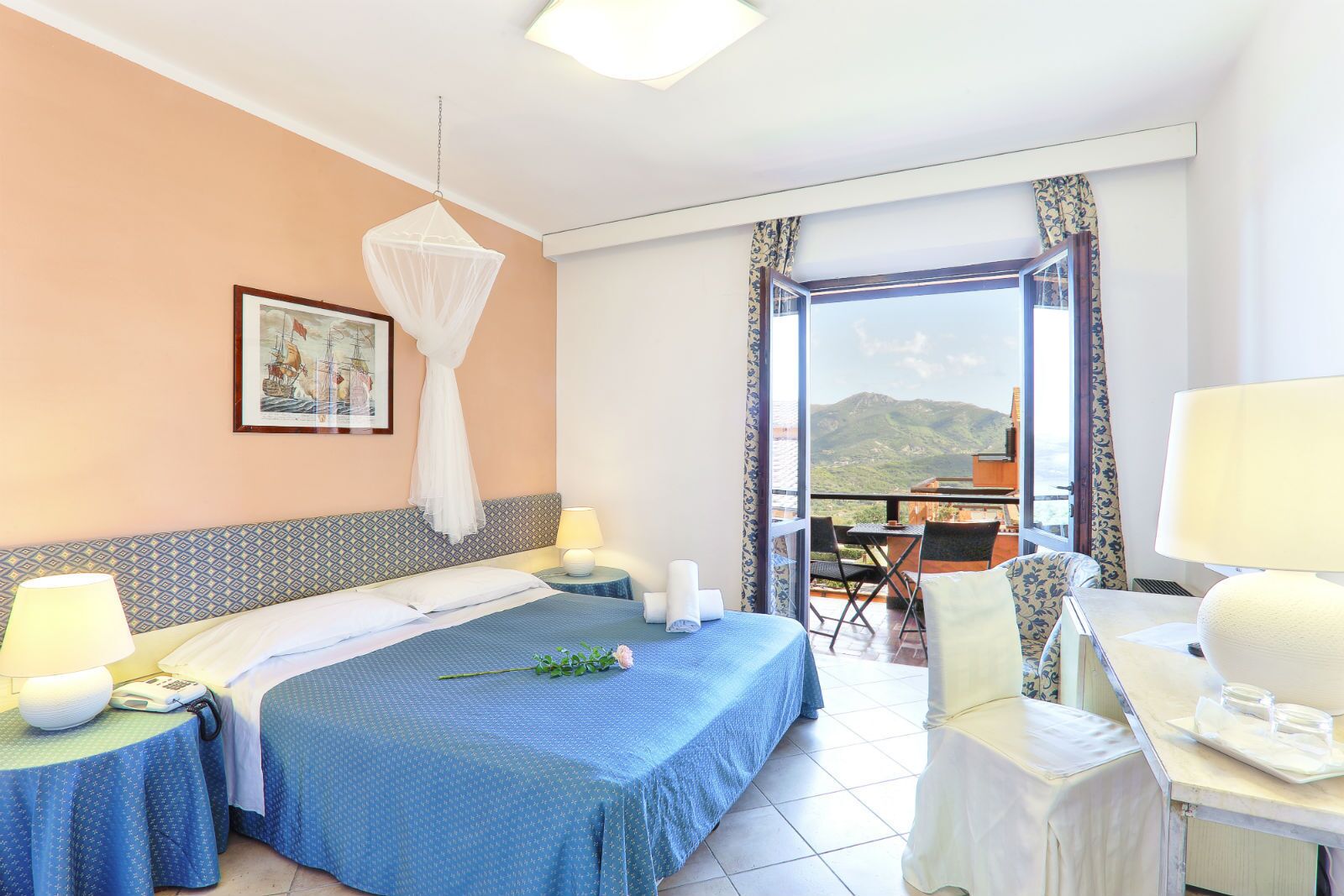 A hotel room with two beds, a balcony with a table and chairs, a telephone, and a painting on the wall.