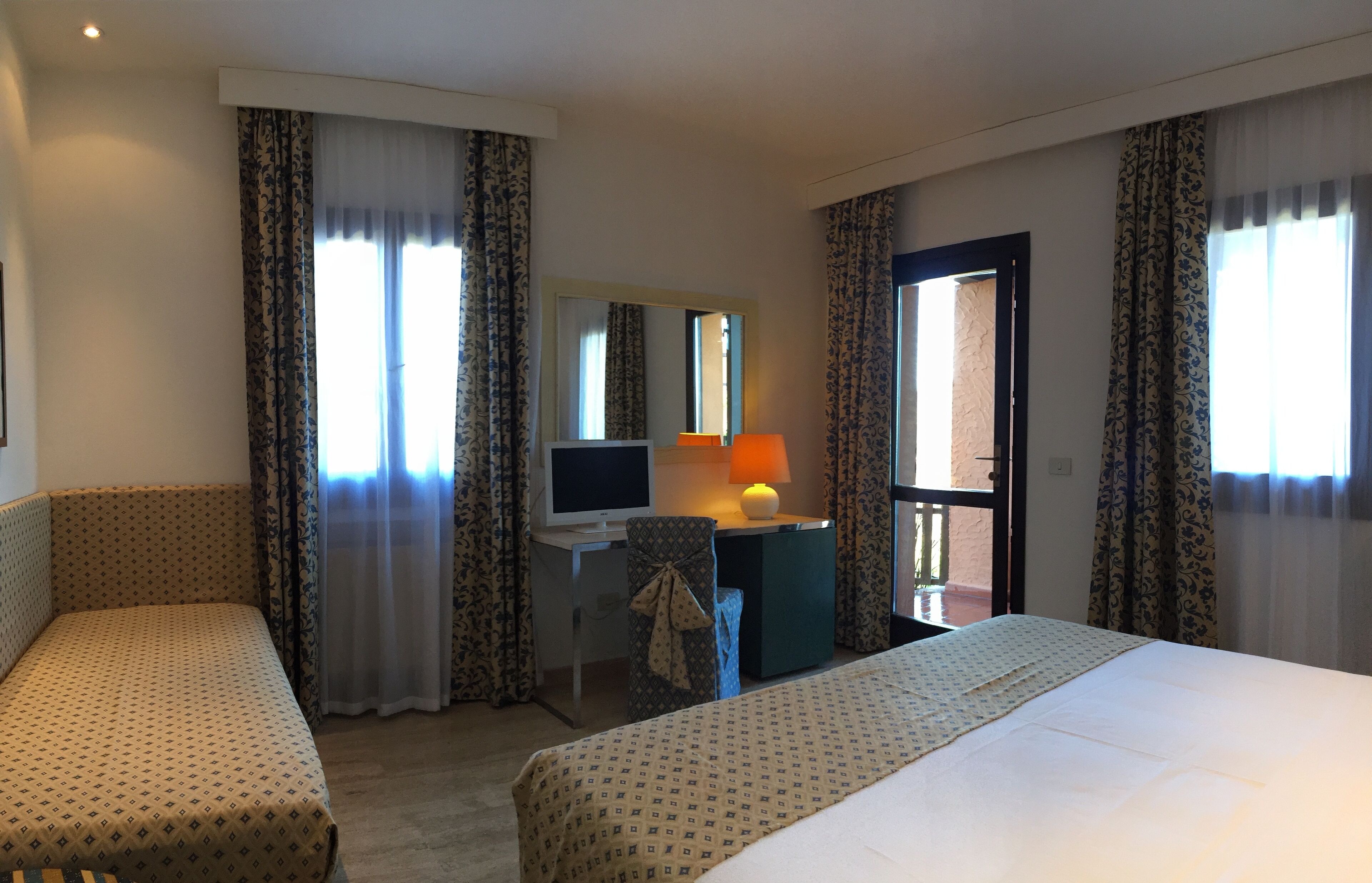 A hotel room with two beds, a desk with a computer, a lamp, and a view of the outside.