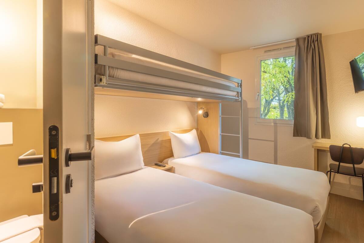 Ein Hotelzimmer mit zwei Betten, einem Schreibtisch, einem Fernseher und einem Fenster mit Vorhängen.