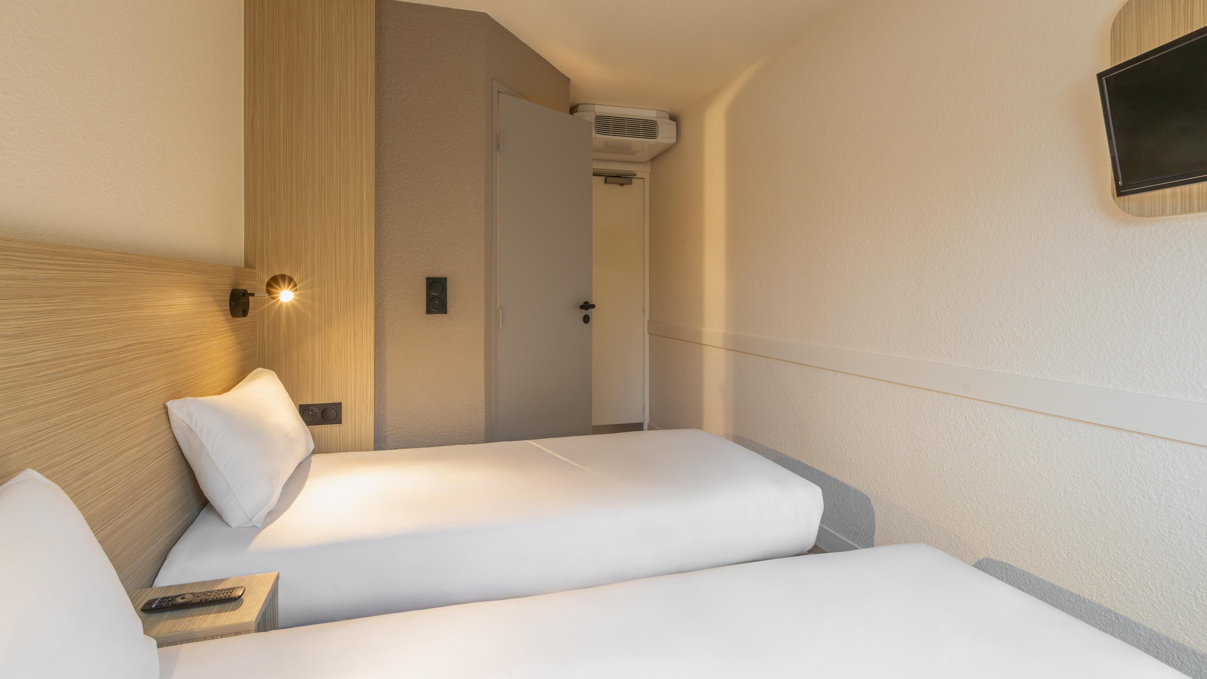 A hotel room with two beds, a wooden headboard, a wall-mounted TV, and a door leading to another room.