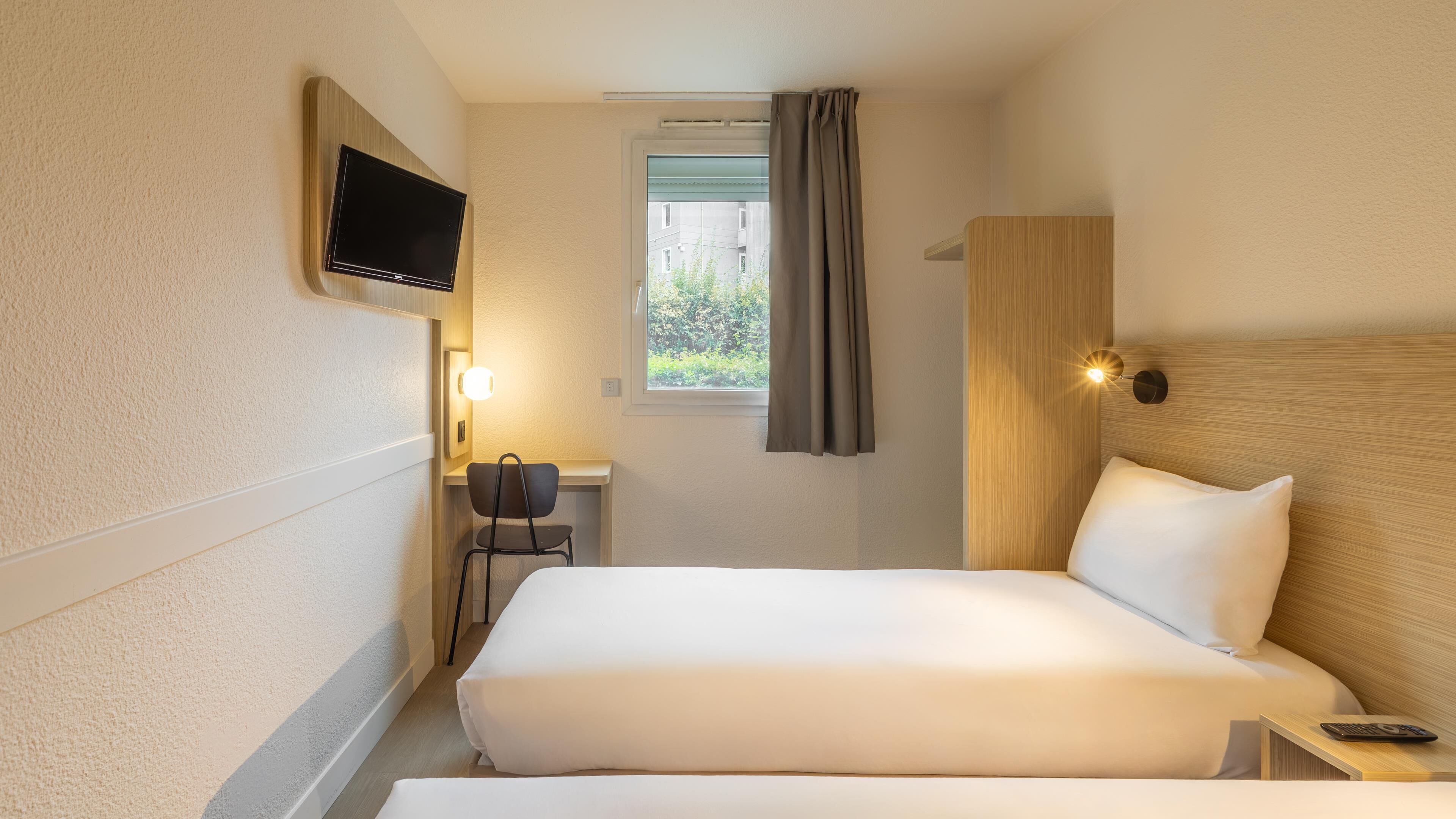 A hotel room with two beds, a desk, a chair, a TV, and a window with curtains.