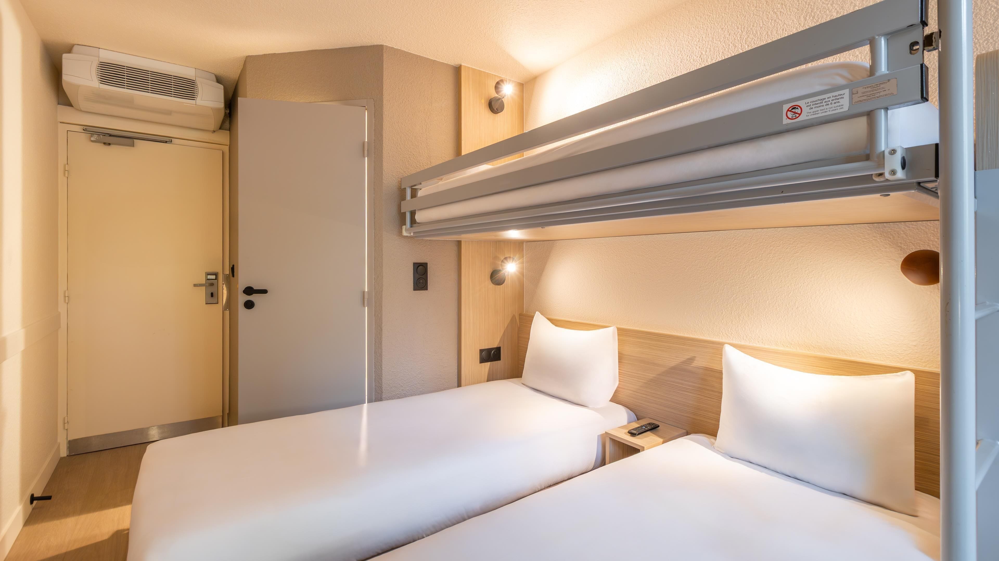 Camera d'albergo compatta con un letto a castello, un letto singolo, un armadio, un riscaldatore e un pannello di controllo.