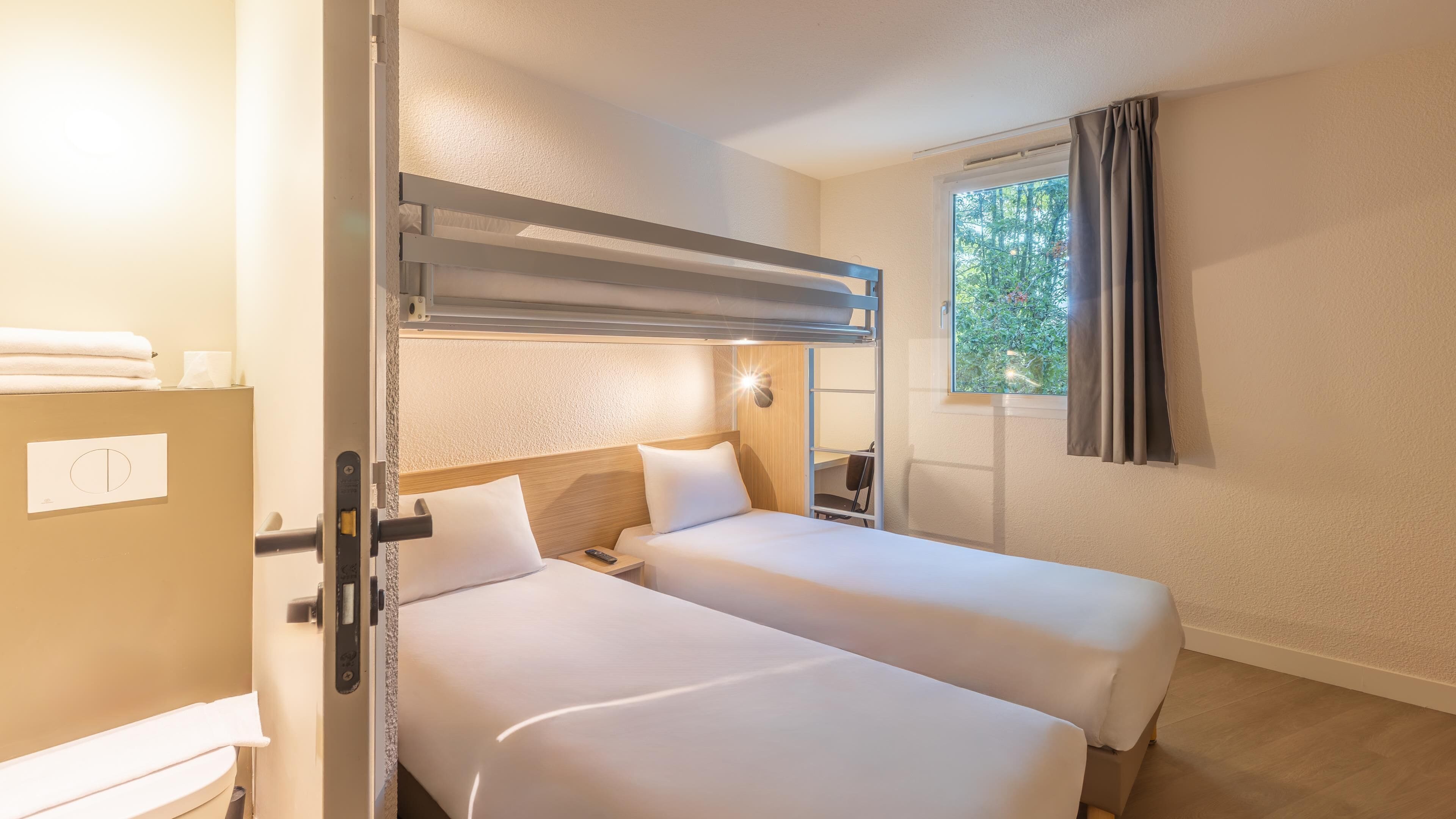 Een hotelkamer met twee bedden, een raam met gordijnen, een nachtkastje met een lamp, en uitzicht op bomen.