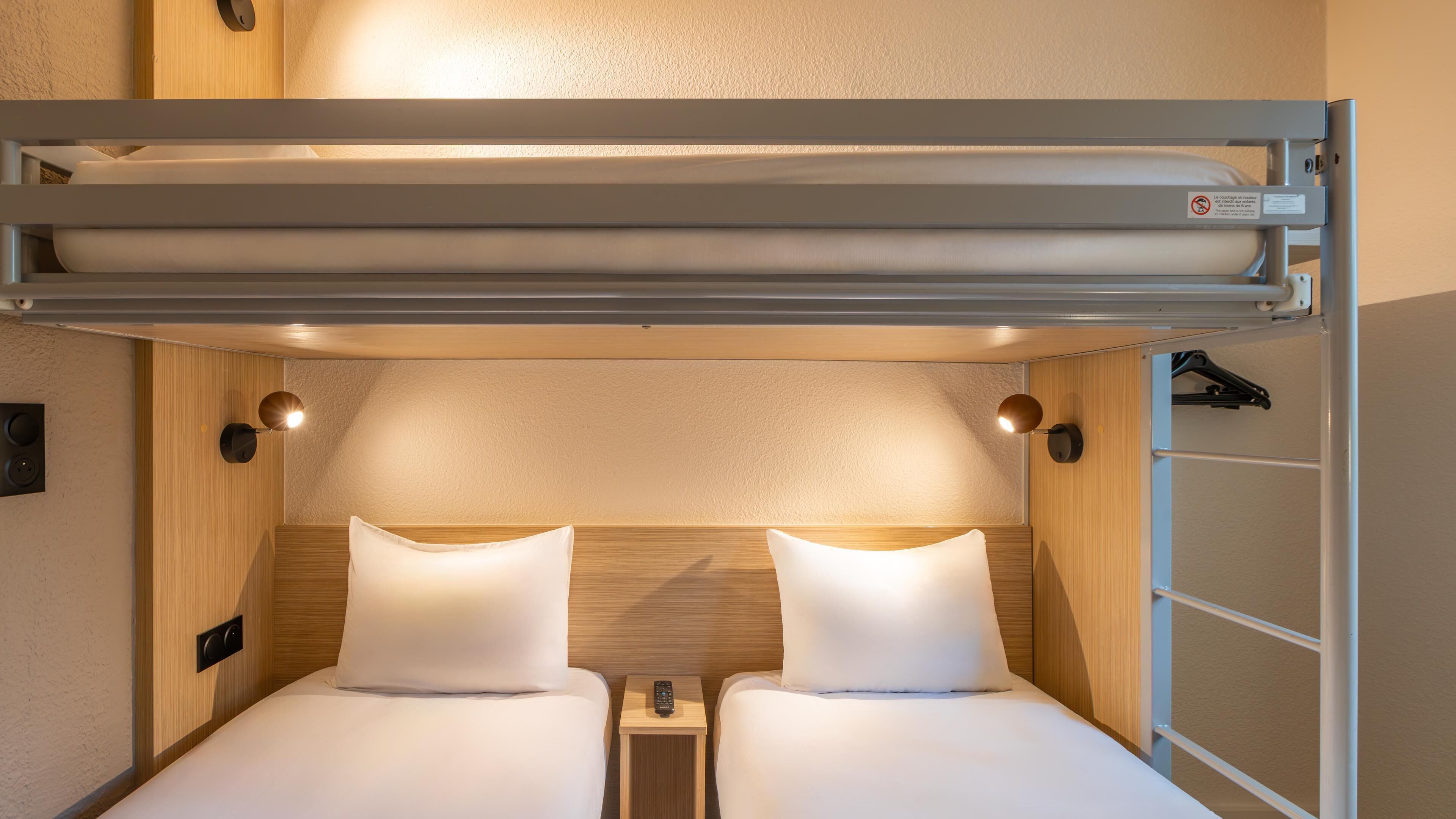 A compact, well-lit room with two beds, a small bedside table, and a built-in bench above the beds.