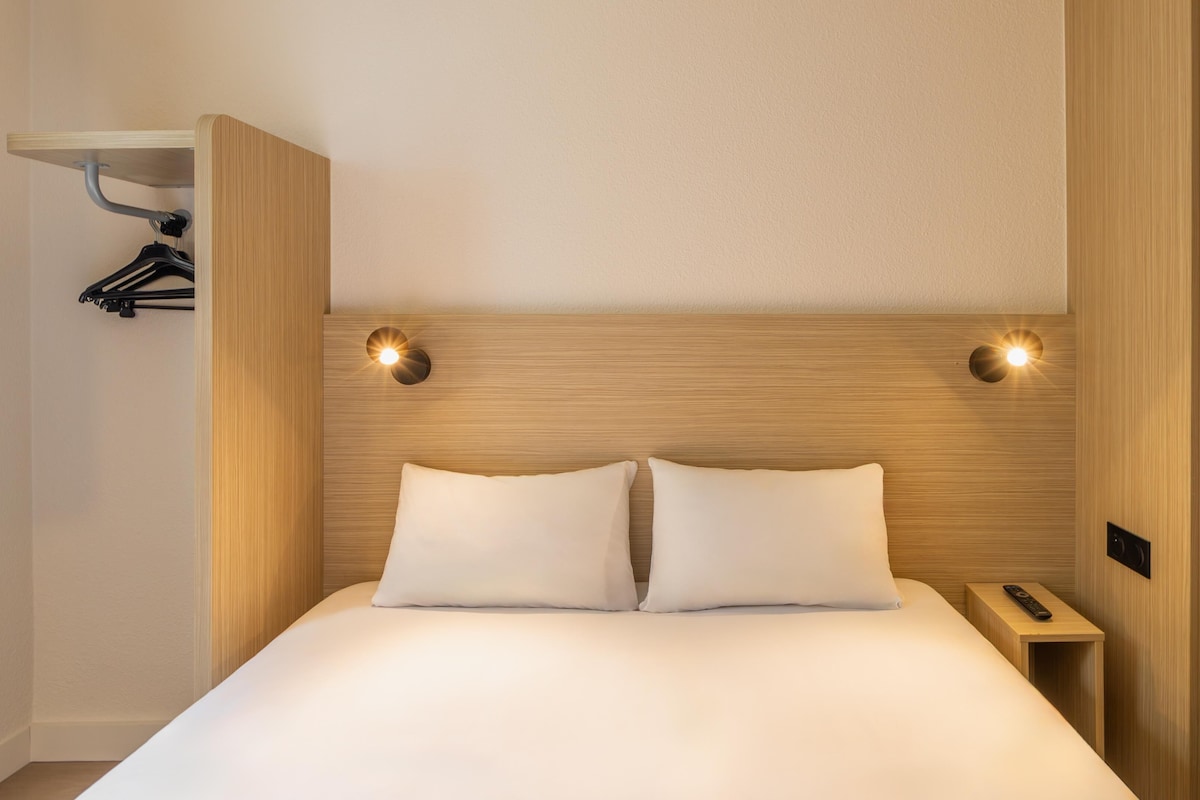 A hotel room with a wooden headboard, two bedside lamps, a bed with white linens, a wooden nightstand, and a coat rack with hangers.