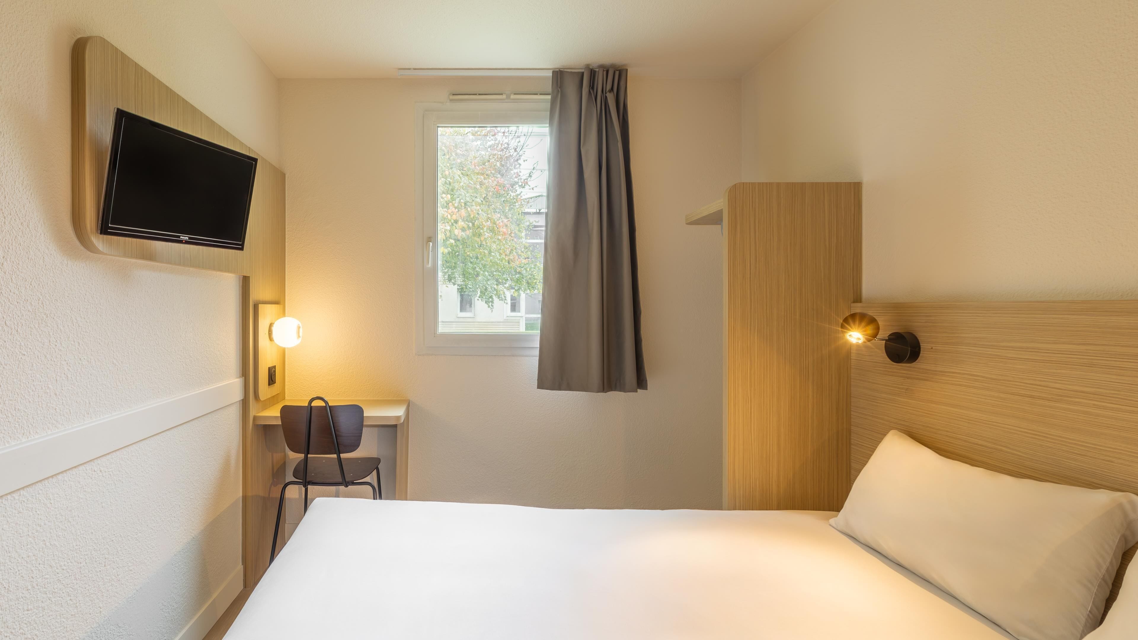 A hotel room with a bed, a desk with a chair, a television, a window with curtains, and a wall-mounted light fixture.
