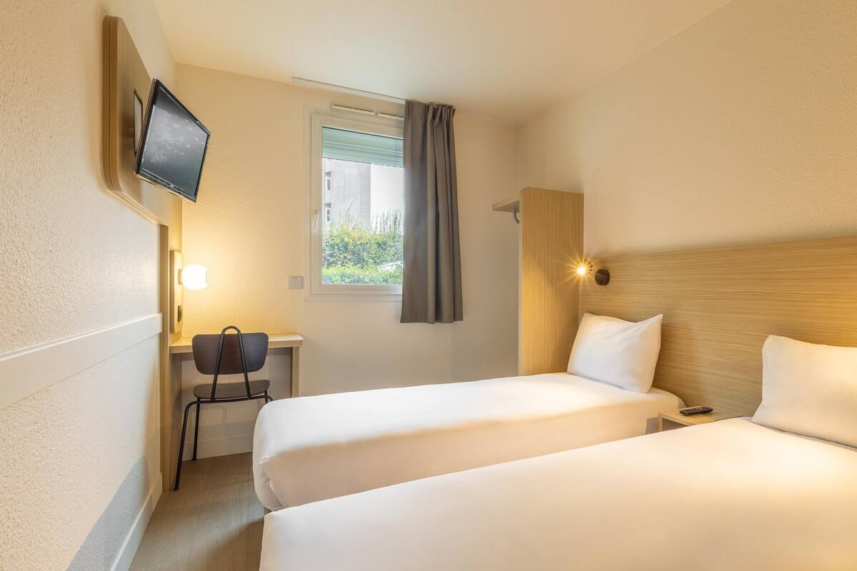 Ein Hotelzimmer mit zwei Betten, einem Schreibtisch mit Stuhl, einem an der Wand montierten Fernseher und einem Fenster mit Vorhängen.