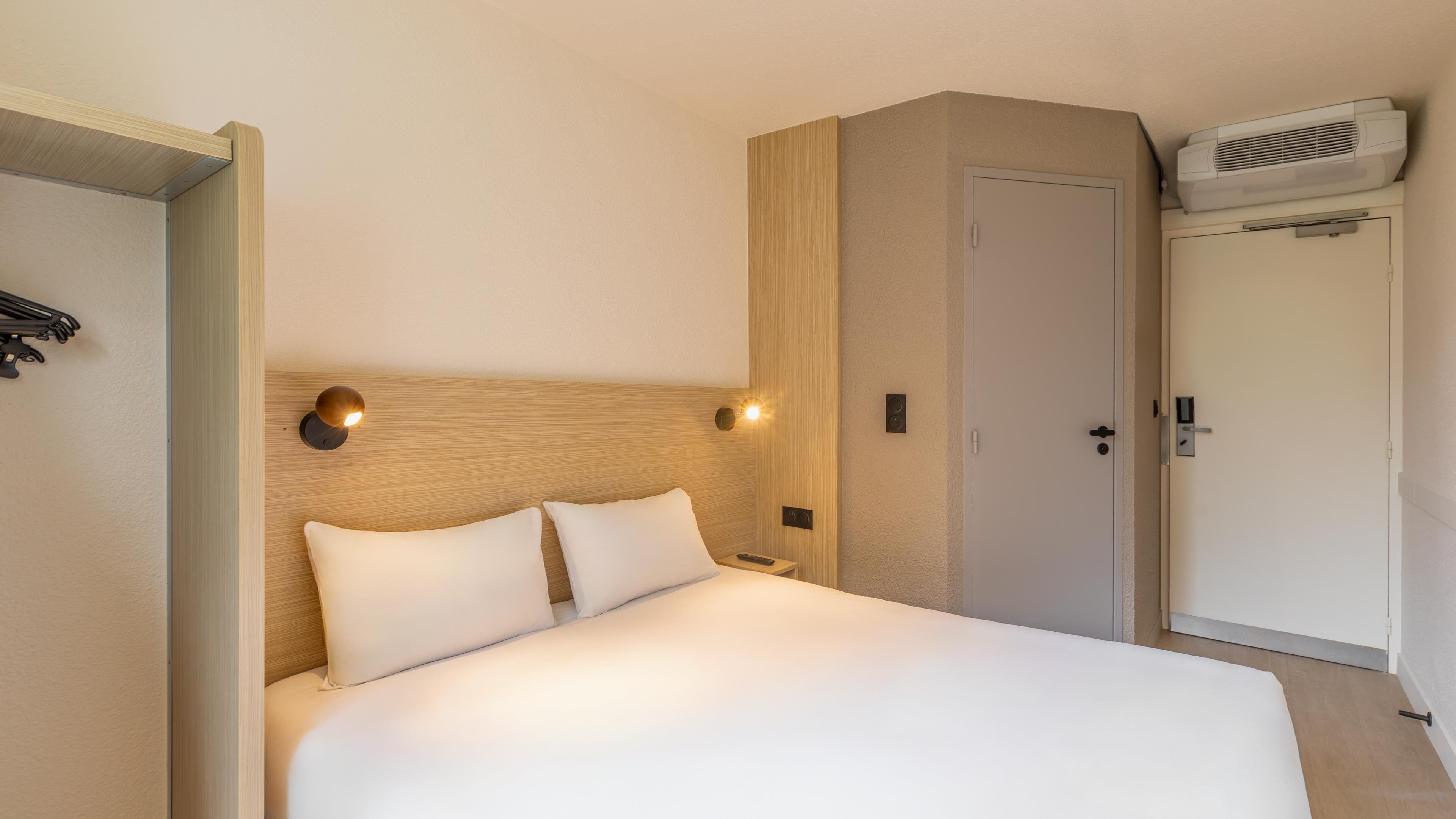 A hotel room with a bed, bedside tables, a wardrobe, and an air conditioning unit.