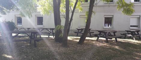 An outdoor seating area with picnic tables, surrounded by trees and a building with windows.