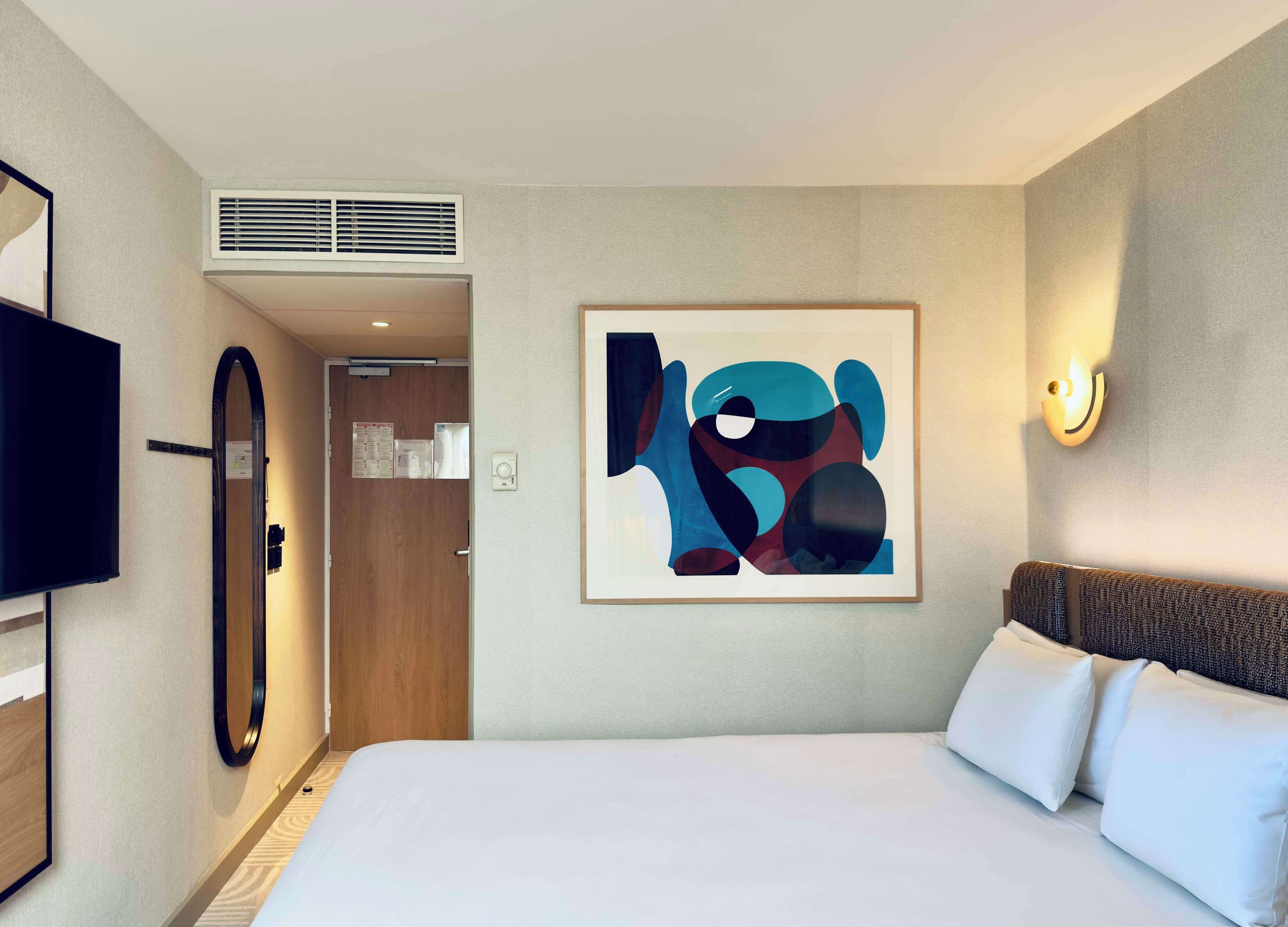 A hotel room with a large bed, a flat-screen TV, a framed abstract painting, a wall-mounted light, and a door with a mirror.