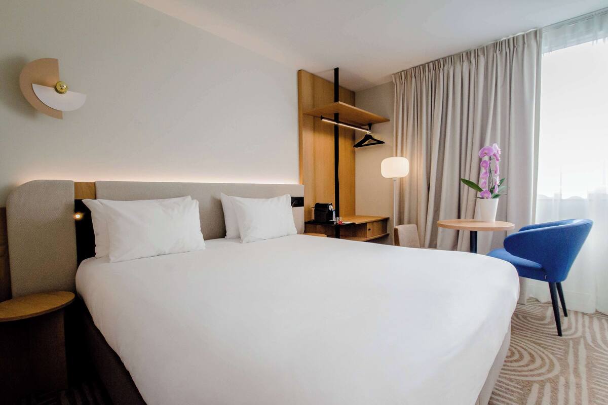 Une chambre d’hôtel avec un grand lit, une table de chevet, une chaise, une petite table, une lampe et une fenêtre avec des rideaux.