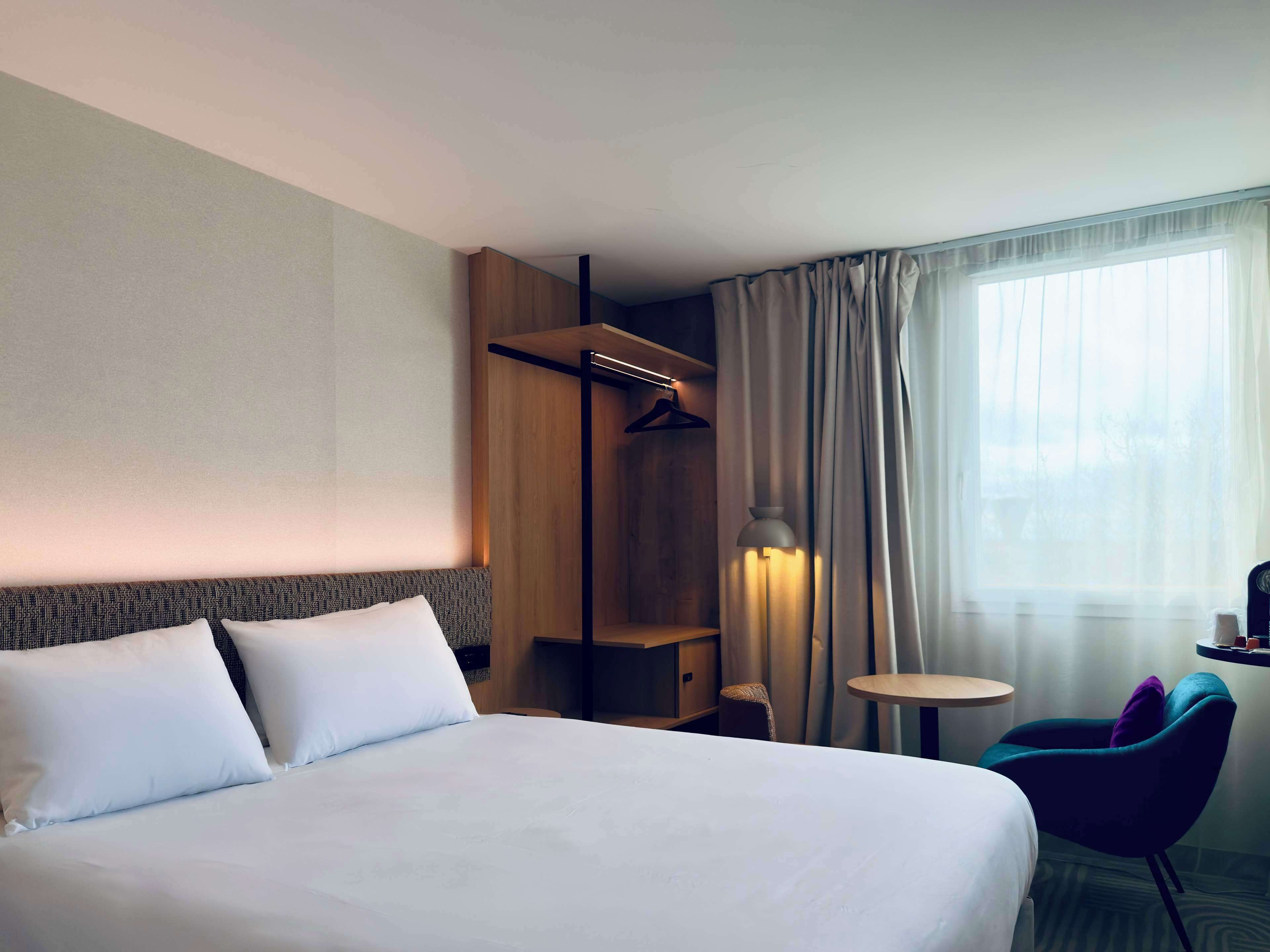 A hotel room with a large bed, a wardrobe, a seating area with a table and chairs, and a window with curtains.