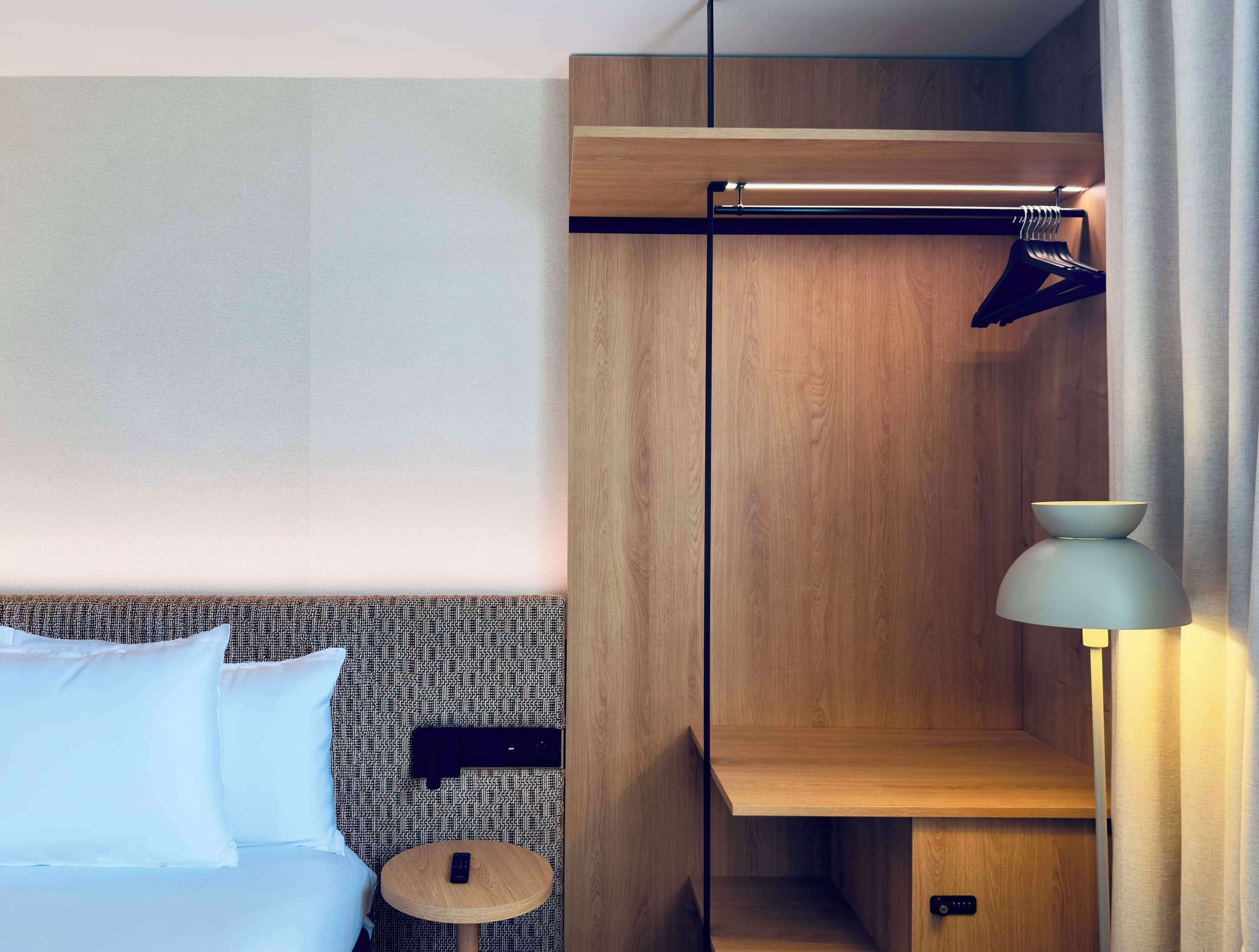 A hotel room with a bed, a small round table, a wall-mounted shelf, and a lamp.