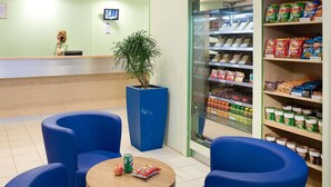 A reception area with a counter, blue chairs, a potted plant, and a refrigerated display case with snacks. - ibis budget Cergy Saint Christophe (Osny)