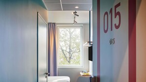A hotel room with a bed, a small table with a chair, a TV, a window with curtains, and a wall with a number. - ibis budget Cergy Saint Christophe (Osny)
