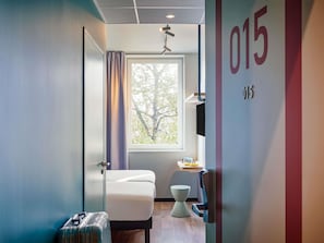 Une chambre d’hôtel avec un lit, une petite table et une chaise, un téléviseur, une fenêtre avec des rideaux et un mur portant un numéro.