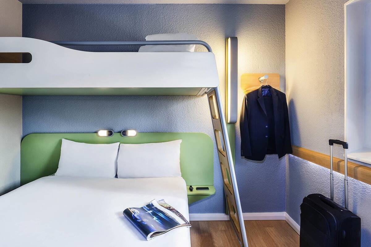 A bunk bed room with a green headboard, a bedside table, a magazine, a suitcase, and a coat hanging on a hook.