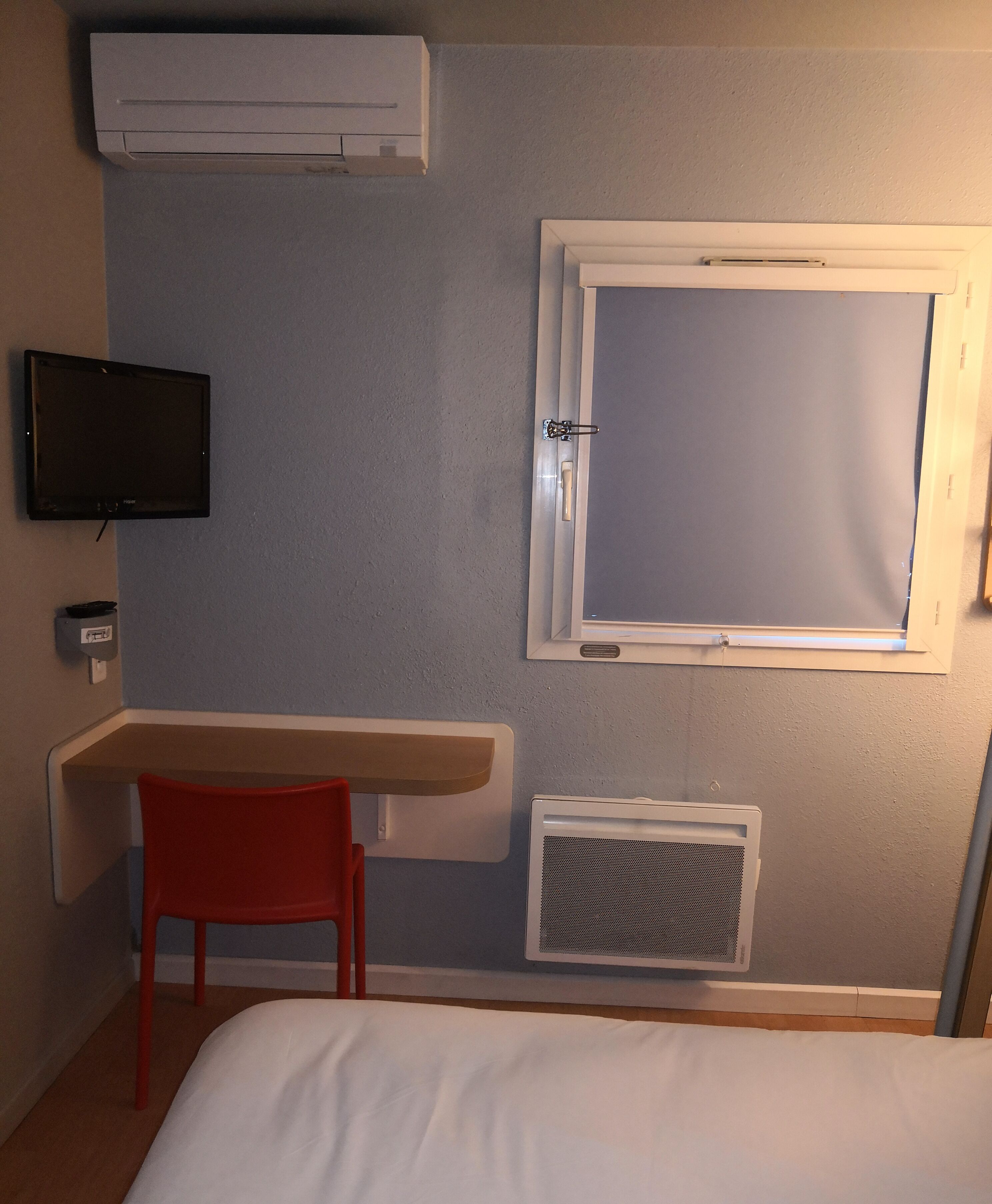 Une chambre comprenant un lit, un bureau avec une chaise, une télévision, une fenêtre et un appareil de climatisation.