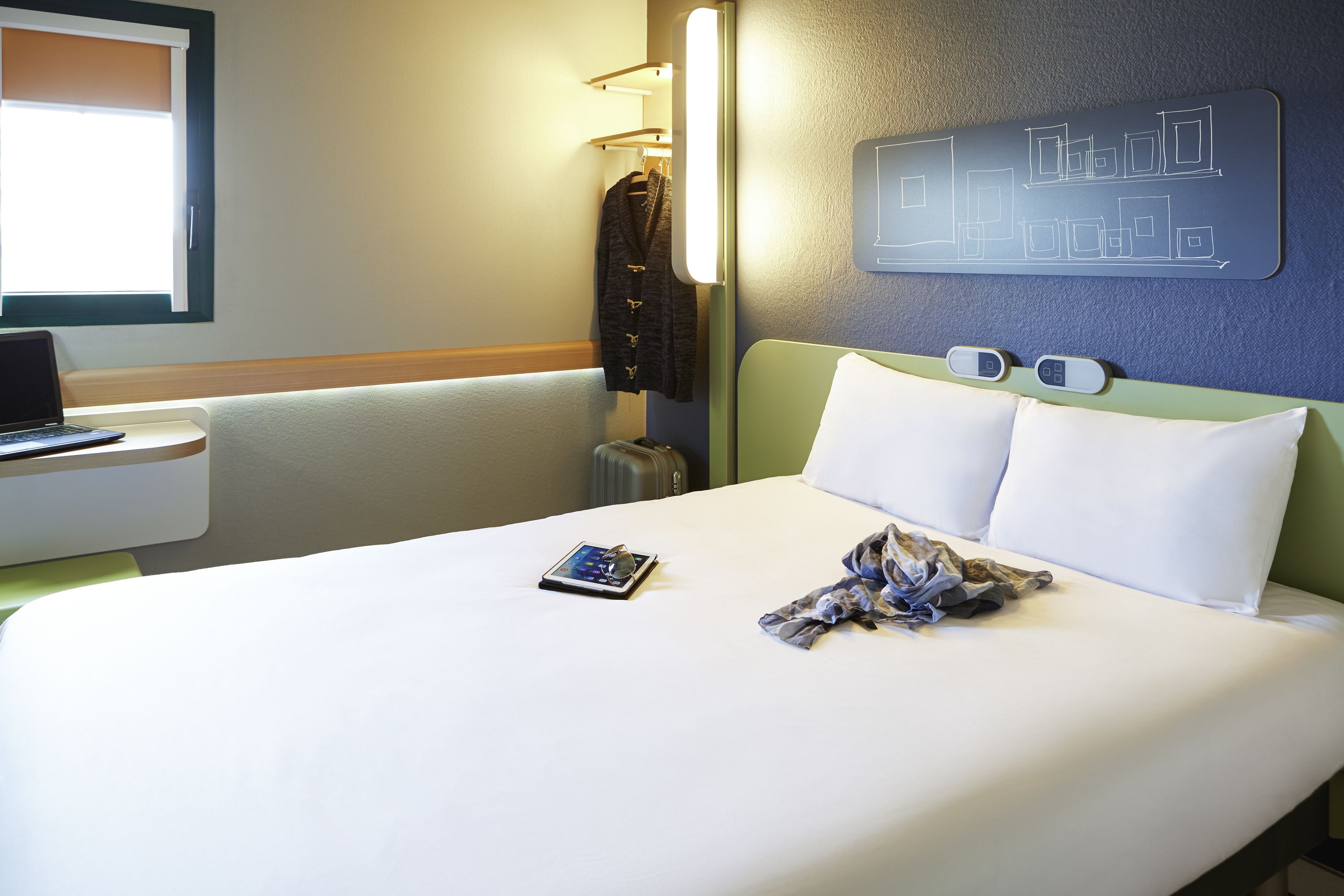 A neatly made bed with white linens, a bedside table with a laptop, and a wall-mounted light fixture.