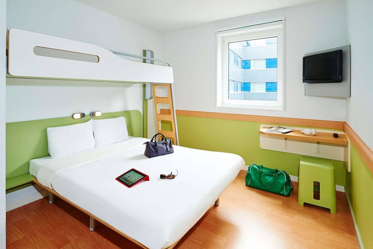 A hotel room with a bunk bed, a bedside table with a lamp, a window with a view of buildings, and a TV mounted on the wall.