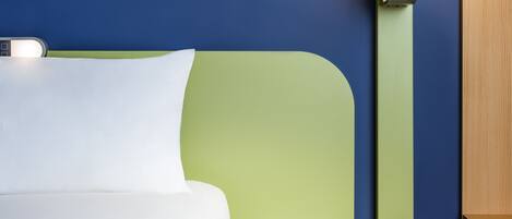 Una cama bien hecha con sábanas blancas, una mesita de noche verde con un libro abierto y un bolígrafo, y una pared azul con un panel de madera.