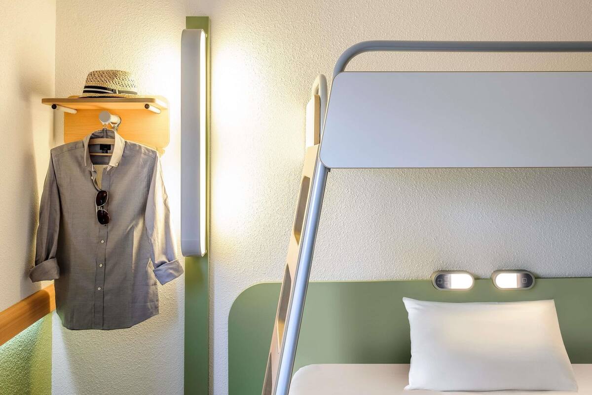 Una cama litera con cabecera verde, un perchero con una gorra y una mesita de noche con una lámpara.