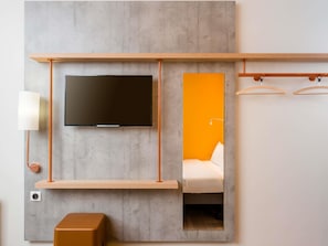 A modern room with a flat-screen TV mounted on a grey wall, a wooden shelf below, a small table, and a bed with an orange accent wall. - ibis budget Cergy Saint Christophe (Osny)