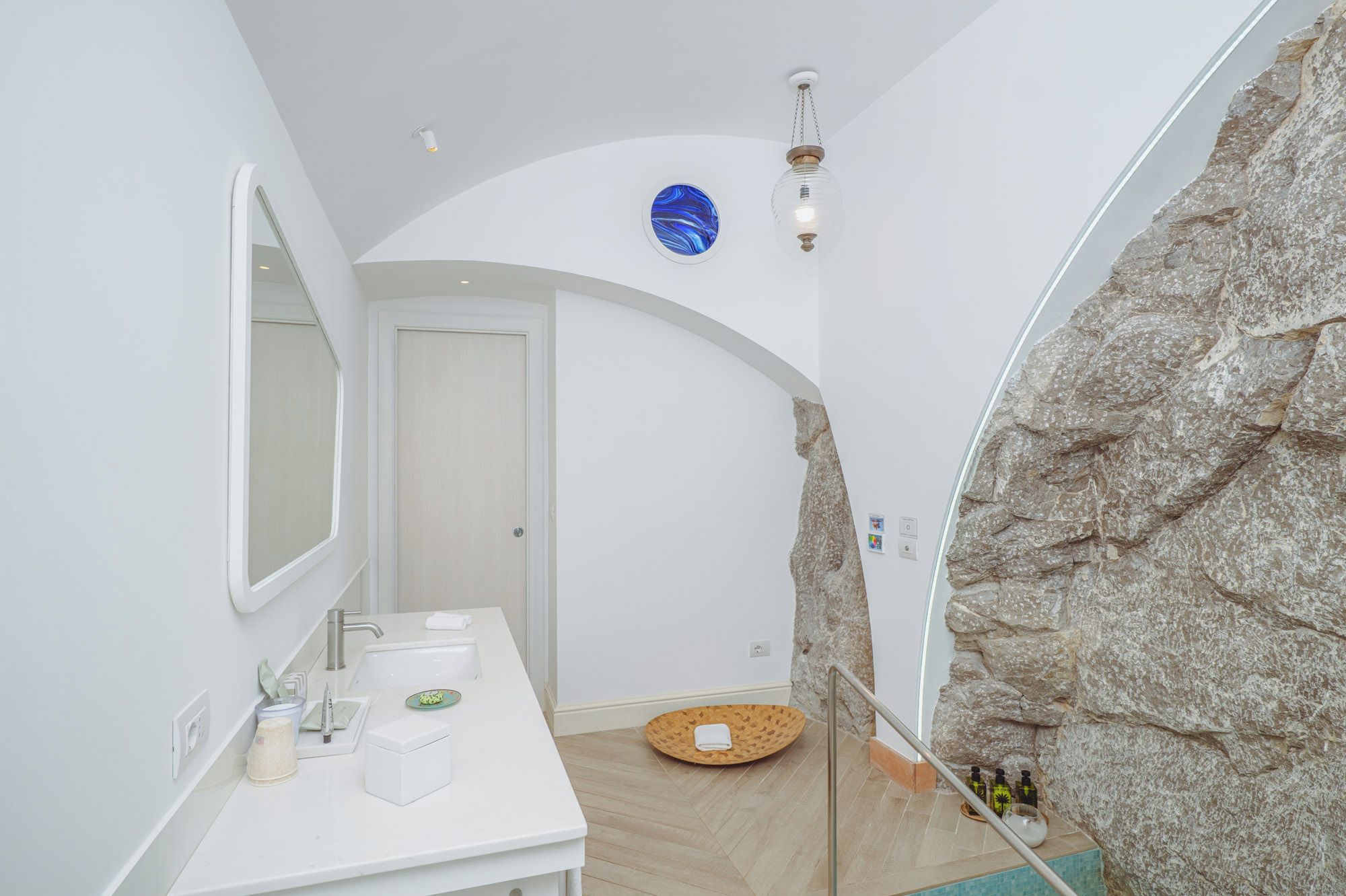 Un baño con tocador blanco, espejo, lavamanos y zona de ducha con pared de piedra.
