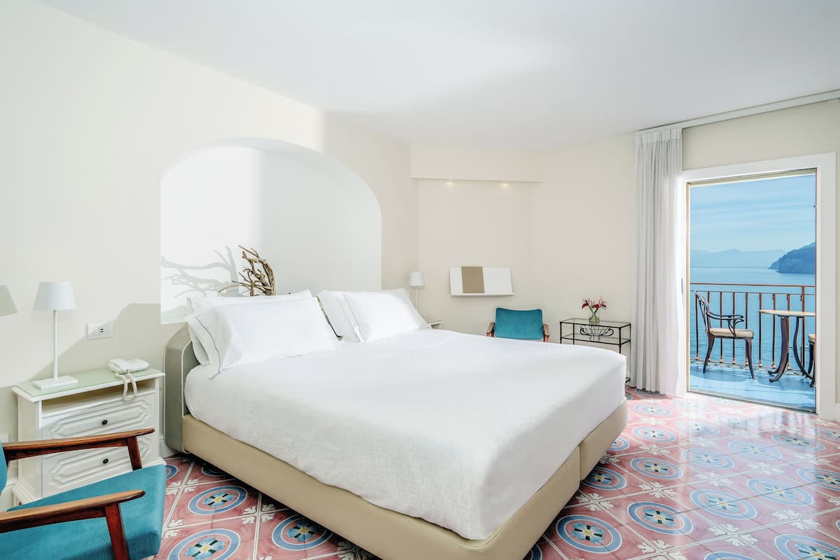 Camera d'albergo con un grande letto, una scrivania, una sedia e un balcone vista mare.