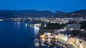 Une vue d’une ville côtière au crépuscule, avec un port de plaisance, des bâtiments et des montagnes en arrière-plan.