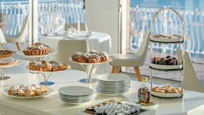 Une table de buffet garnie de diverses pâtisseries et desserts, une bouteille de sirop et un présentoir à étages sur lequel sont disposées d’autres pâtisseries. La scène se déroule dans une pièce lumineuse, dotée de grandes fenêtres et offrant une vue sur la mer.