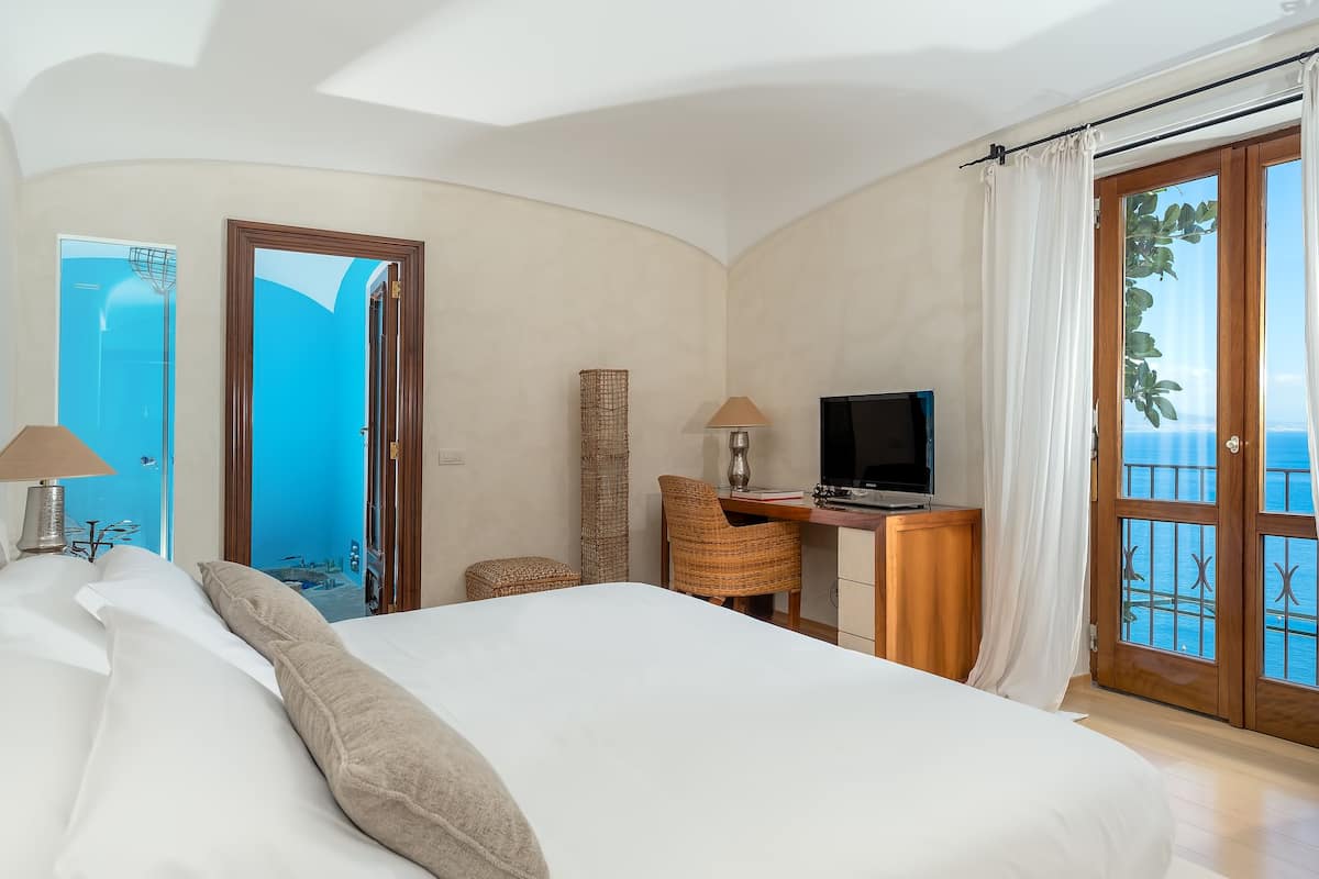 Camera da letto con un letto grande, una scrivania con TV, una sedia e vista mare tramite una porta in vetro.