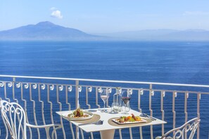 Uma mesa com pratos de comida e copos de vinho numa varanda com vista para o mar.