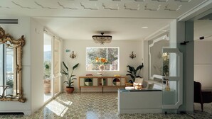 A spacious room with patterned floor tiles, a large ornate mirror, a wooden console table with decorative items, and a large window with a view of greenery.