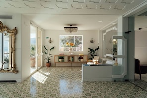 A spacious room with patterned floor tiles, a large ornate mirror, a wooden console table with decorative items, and a large window with a view of greenery.