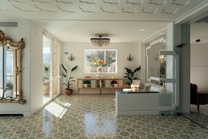 Una habitación amplia con baldosas de diseño, un espejo grande y ornamentado, una consola de madera con objetos decorativos y un ventanal con vistas a zonas verdes.