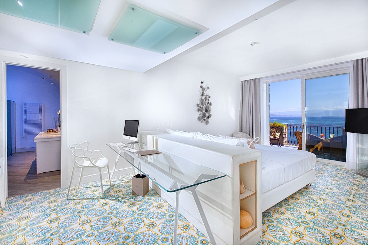 Una moderna camera d'albergo con una scrivania in vetro, un letto e un balcone vista mare.
