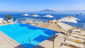 A pool area with lounge chairs and umbrellas, overlooking the sea with a distant mountain view.