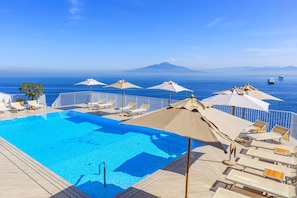 A pool area with lounge chairs and umbrellas, overlooking the sea with a distant mountain view.