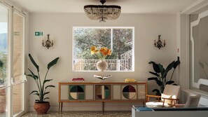 Quarto com uma grande janela, um candelabro, uma mesa consola com livros e plantas em vasos.