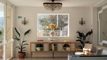 Una habitación con un ventanal grande, una araña, una consola con libros y plantas en macetas.