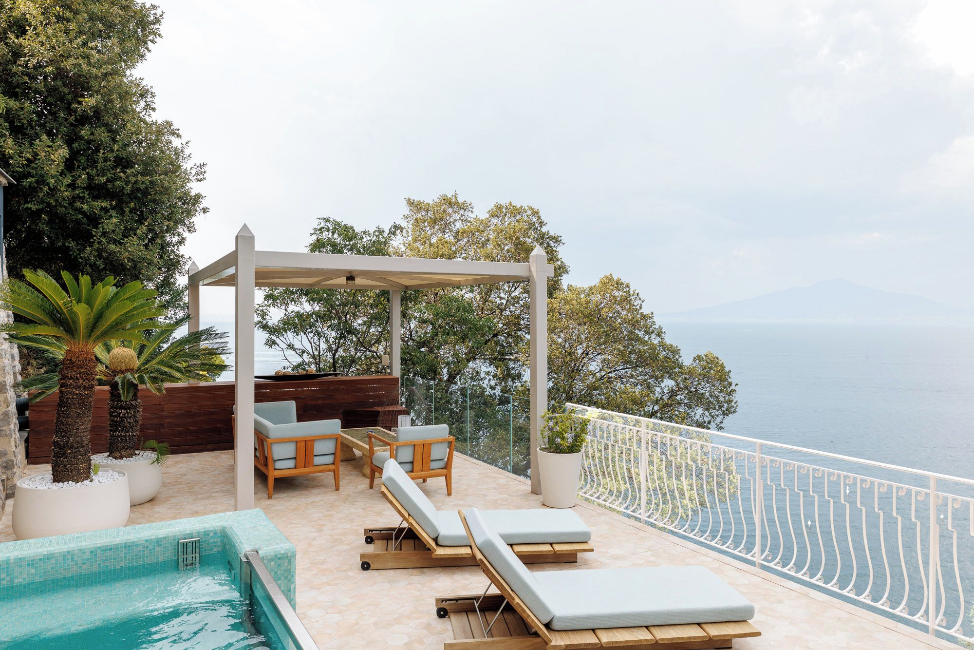 A poolside area with lounge chairs, a pergola, and a view of the sea.